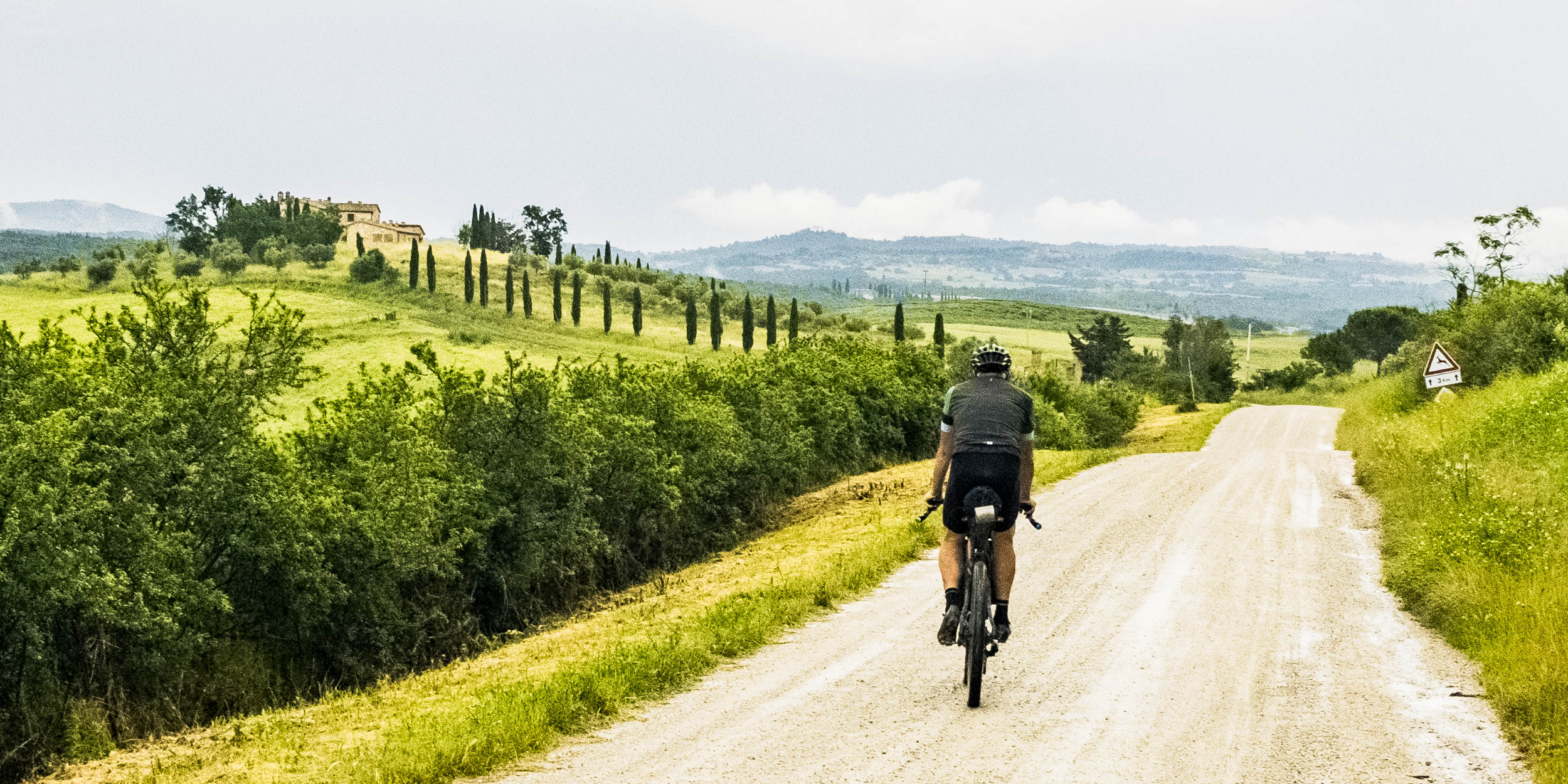 Radfahrer mit Gravelbike auf toskanischer Strade Bianche mit Hügellandschaft und Zypressen im Hintergrund